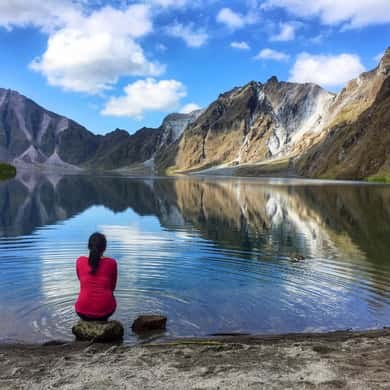 A visitor sitting on a boulder at the water's edge with the crater rim mirrored behind
